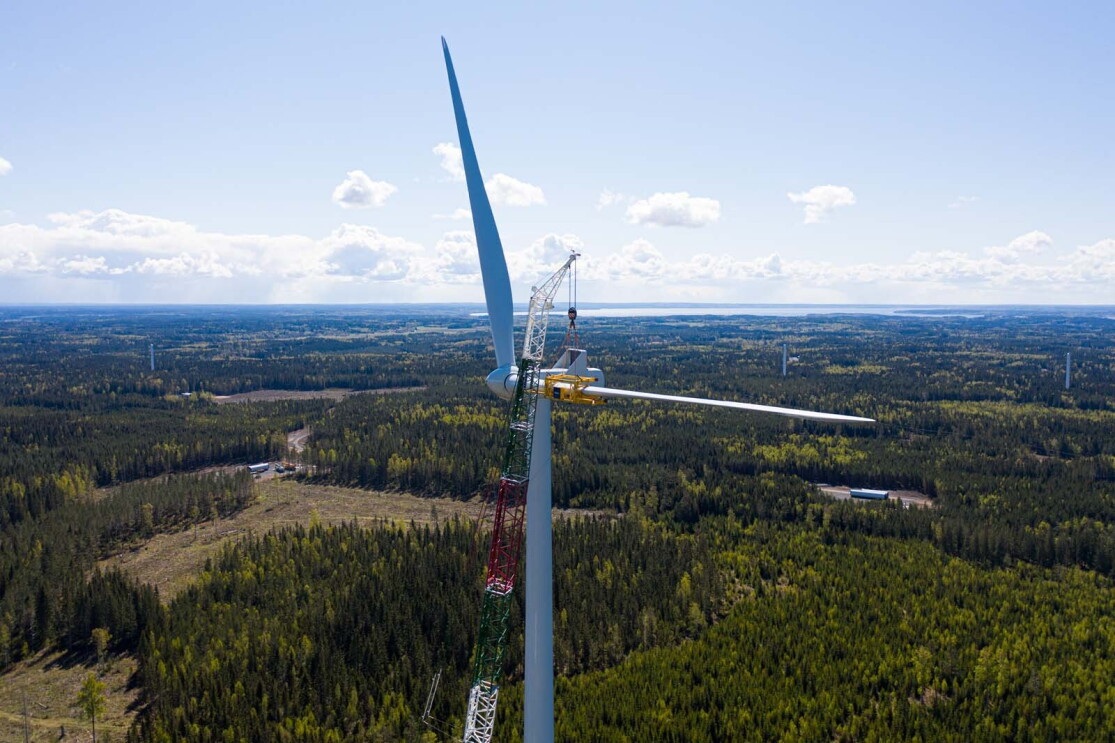 Aerial view photo of a crane assembling a wind turbine with the horizon as a backdrop, in Bäckhammar, Sweden.
