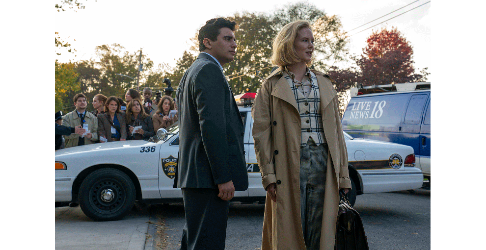 規制テープが張られた屋外で、スーツ姿の人物とトレンチコートの人物が立っている。2人の背後にパトカーと報道車両、手にメモを持った報道陣。