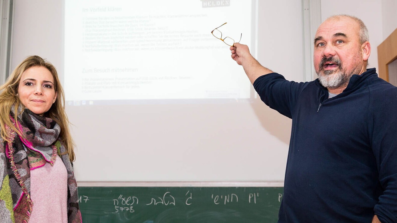 Ein Mann und eine Frau stehen vor einer Wand, auf der eine Präsentation läuft. Sie schauen in die Kamera. Der Mann zeigt mit seiner Hand auf die Präsentation hinter sich.