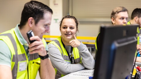 Amazon Logistik Mitarbeiter im FC, sie tragen gelbe Sicherheitswesten, sie stehen am Schreibtisch vor Bildschirmene und unterhalten sich