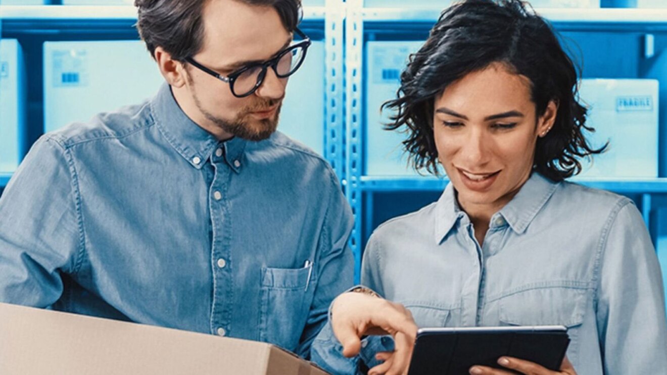 En una foto editada a nivel de color, aparece un hombre y una mujer, los dos con una camisa tejana y mirando la tablet que sujeta la mujer. El hombre aparece a la izquierda de la imagen, sujeta un portátil con la mano derecha y con la izquierda señala la tablet de la mujer, Lleva el pelo corto, barba de dos días y unas gafas negras de pasta. La mujer tiene el pelo ondulado. Pendientes y con la mano izquierda sujeta la tablet. De fondo hay estanterías con material. Solo los protagonistas están en los colores originales, el resto de la foto es de color azul.