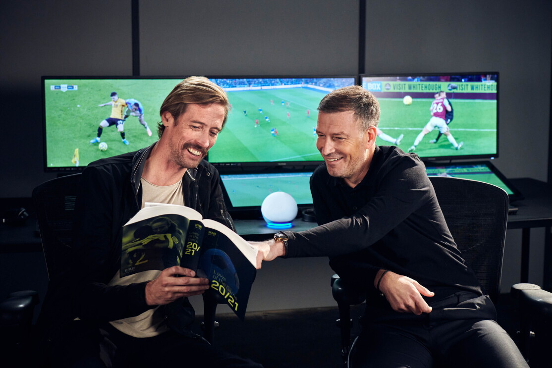 Peter Crouch (left) and Mark Clattenburg (right) reading through a football rule book in front of screens showing football games. On a table between them is the Amazon Echo Dot.