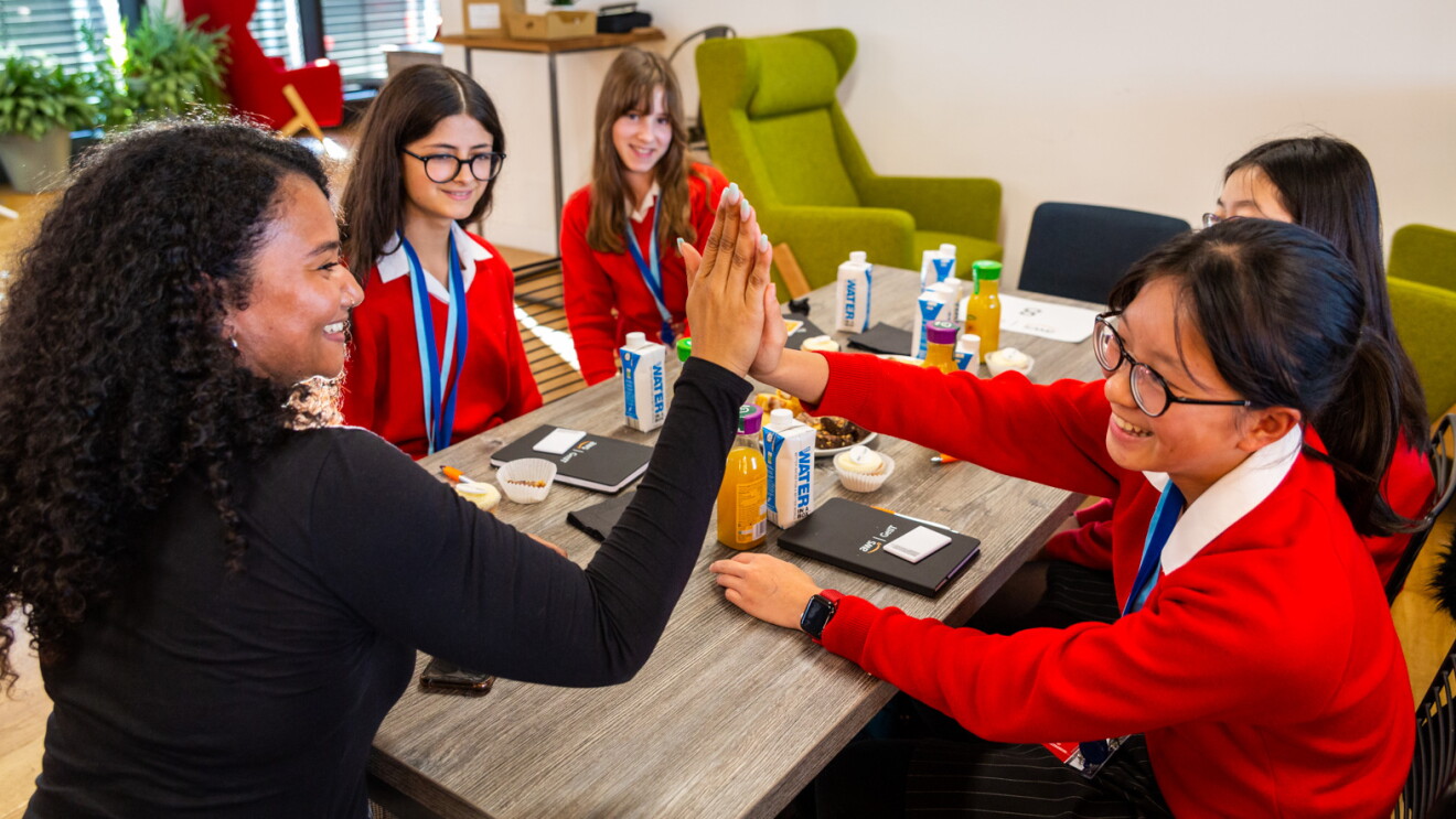 AWSGetIT. En la foto hay cuatro chicas vesttidas con uniforme: camisa blanca, jersey rojo y un collar de dos colores azules. Hay también una señora con el pelo rizado negro, y un jersey negro. Las cinco mujeres etán sentadas en una mesa.  y dos de ellas están chocando las manos. Encima de la mesa libretas negras con las letras de AWS y aguas y zumos. De fondo un par de sofás y una pequeña mesa.