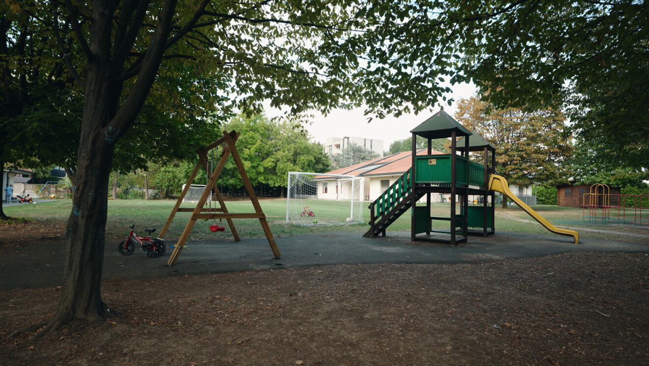 Aldea de Aldeas Infantiles SOS de Italia y desde la aldea de Saronno. Unos columpios de madera y una construcción de dos casas de madera con un tobogán de color amarillo. De fondo un campo de fútbol y dos porterías y unas viviendas.  