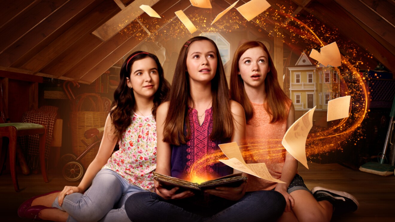 Three girls sit together. One holds a glowing book with pages flying out of it.