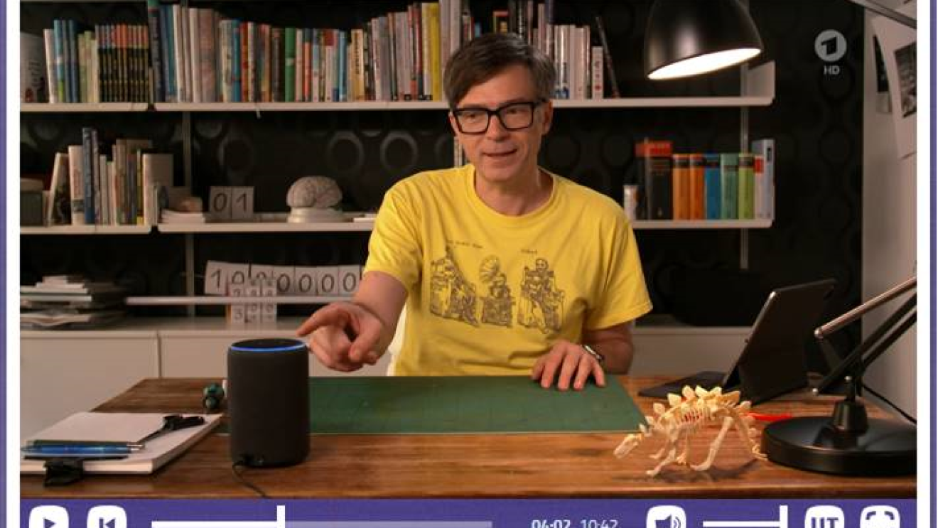 Ein Mann in einem gelben Tshirt sitzt an einem Schreibtisch und zeigt mit dem Finger auf ein Alexa Echo.
