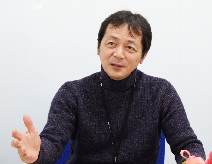 白い壁の前で両手を広げて話す男性。黒に近いグレーのニットを着ている。