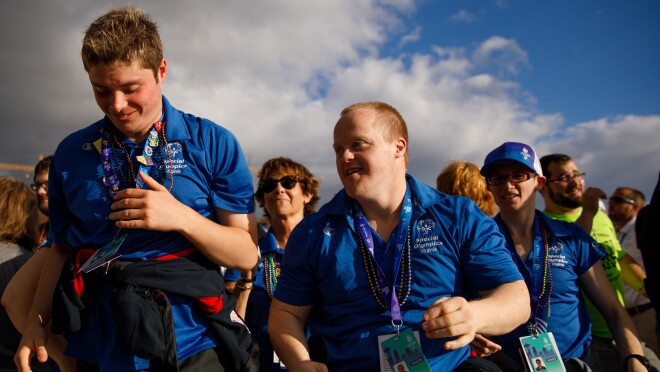 来自弗吉尼亚州的特奥会运动员和与会者似乎是一群人的领导者。前景中有两只微笑的雄性,似乎正在向前移动。在中间地带,另外两名穿着“弗吉尼亚特奥会”t恤的人面向同一个方向。在背景中,可以看到更多的与会者。