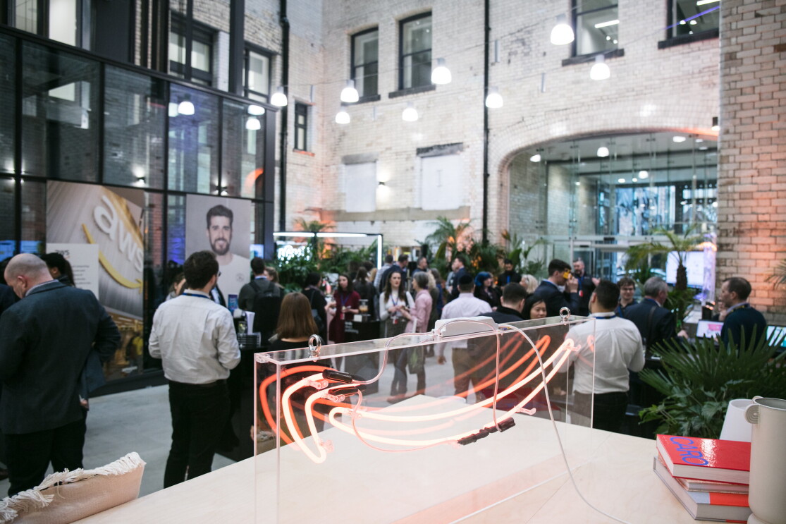 A photo of Amazon employees standing and chatting at the Manchester Corporate Office opening. On a table, there is a large Amazon smile logo. 