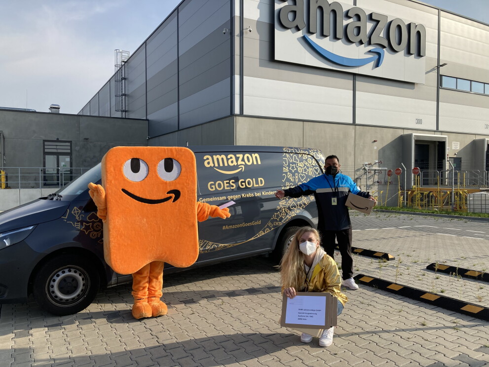 Amazon Maskottchen Peccy, eine Mitarbeiterin und ein Lieferpartner stehen vor einem mit "Amazon Goes Gold" beschrifteten Liefervan. Im Hintergrund ist ein Amazon Verteilzentrum zu sehen.