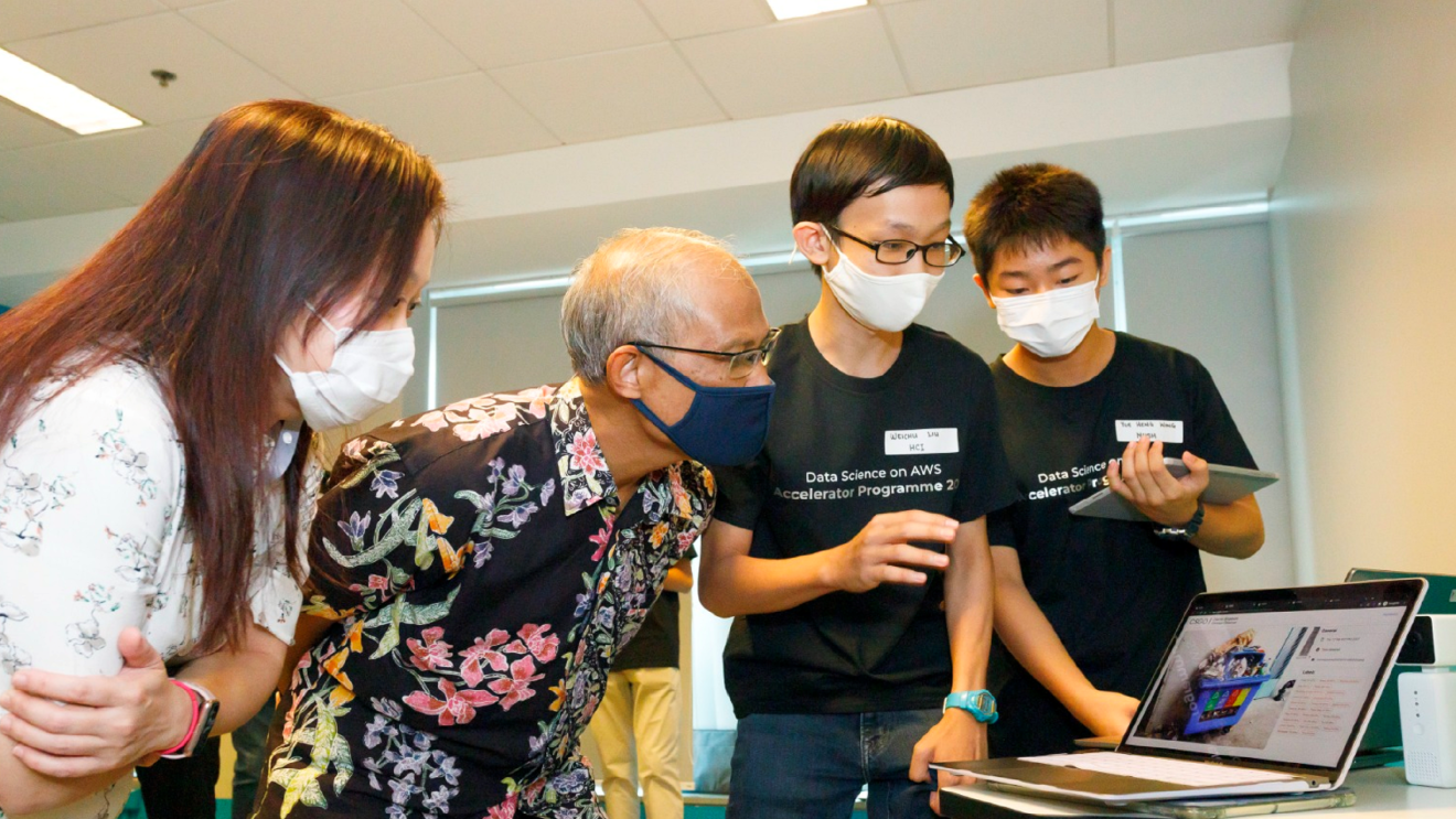 A man in a flora shirt examines the work presented by two teenagers in black t-shirts while a woman in a white dress looks on.