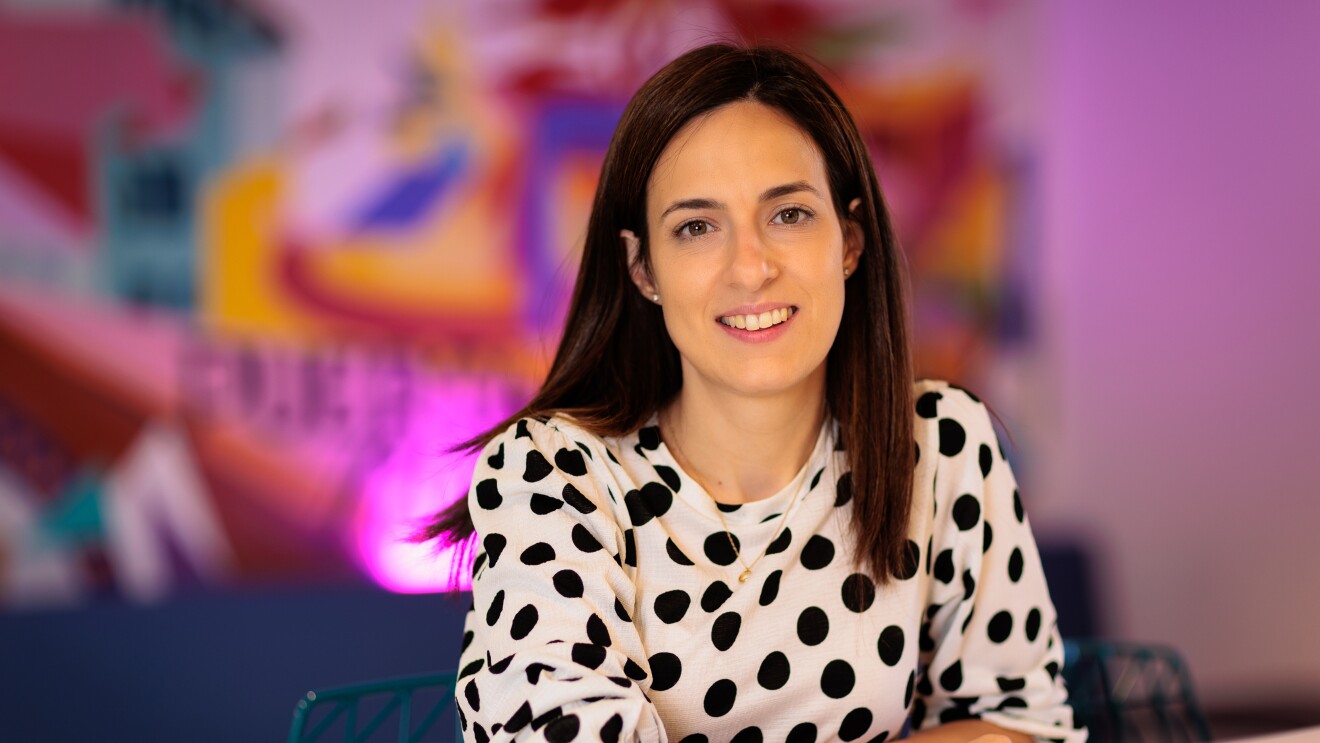 Alejandra Ossa tiene el pelo negro largo hasta el hombro. Lleva un vestido blanco de lunares negros de manga. Está apoyada en la mesa con los brazos cruzados. Fondo de muchos colores vivos difuminados. lleva pendientes, collar y pulsera.