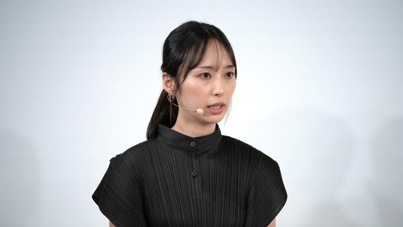 後ろに結んだ髪型をしている黒いブラウスを着ている女性が右に向かって、ヘッドセットマイクで話している(高山芽衣氏)