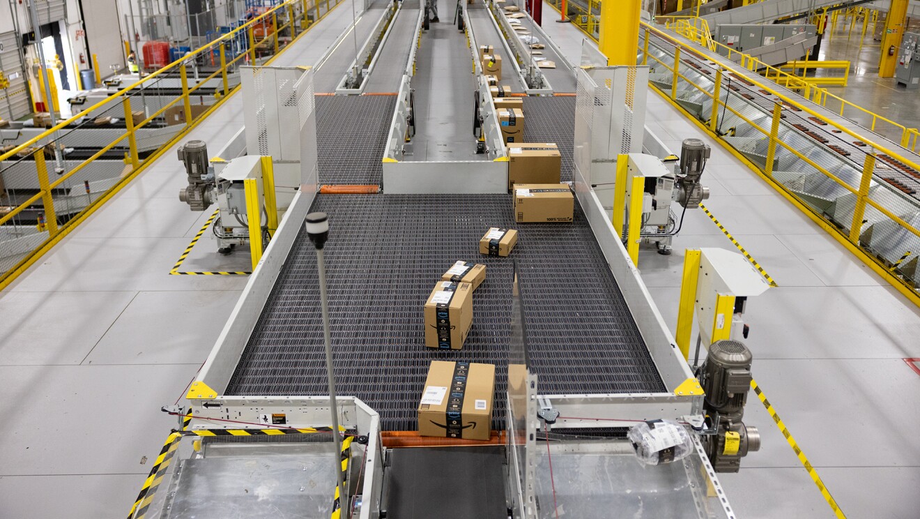 Panorámica de un centro logístico de Amazon. Aparece la parte superior del centro, una cinta transportadora de pedidos. La cinta está llena de cajas. La estructura metálica es de color amarillo. En la parte inferior se ven más cintas transportadoras.
