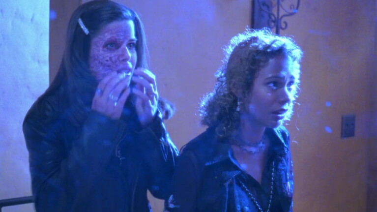 Two women in dark clothing under blue lighting, one with straight hair eating