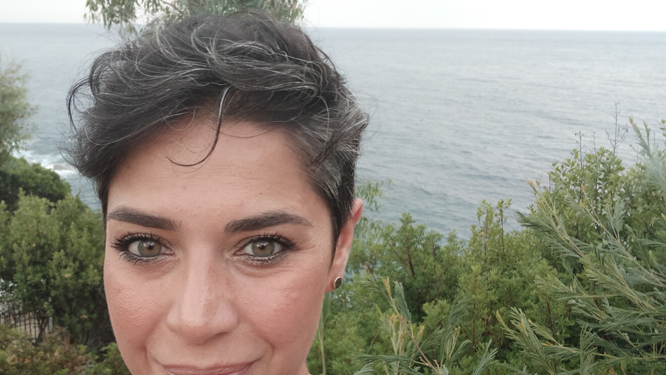 Primo piano di Alessandra, volontaria Amazon. La donna ha i capelli corti e posa sullo sfondo di un ambiente naturale, con il mare alle spalle. 