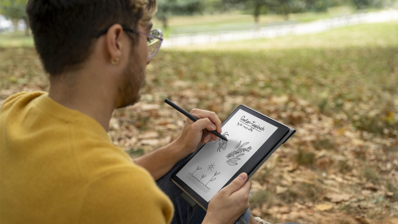 Ein Mann mit Brille und gelben Pullover sitzt in einem Park. Er hält den neuen Kindle Scribe in der Hand, mit welchem er Bilder in sein "Garten Tagebuch" schreibt.