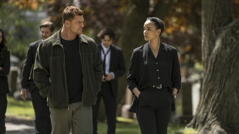 Still from Prime Video series 'Neagley' of man in olive jacket and woman in black suit walking together outdoors in conversation