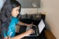 A young woman types on a laptop at a desk in her home. She wears a blue t-shirt from a coding program.