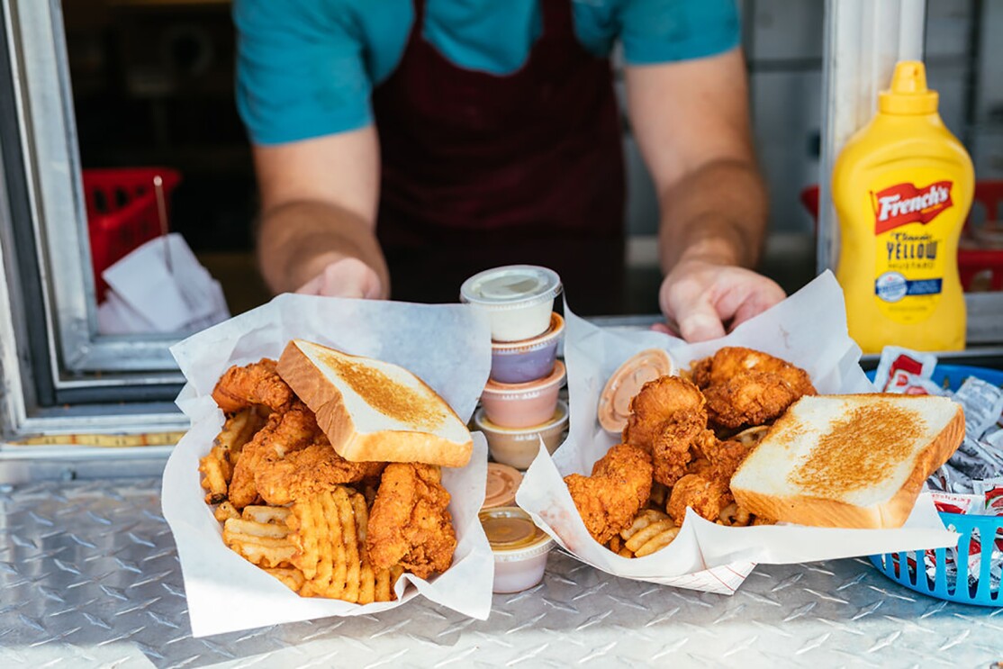 Baskets of chicken fingers, waffle fries and Texas toast are passed through the window of a food truck. A stack of condiments and sauces balances between the two baskets of food.