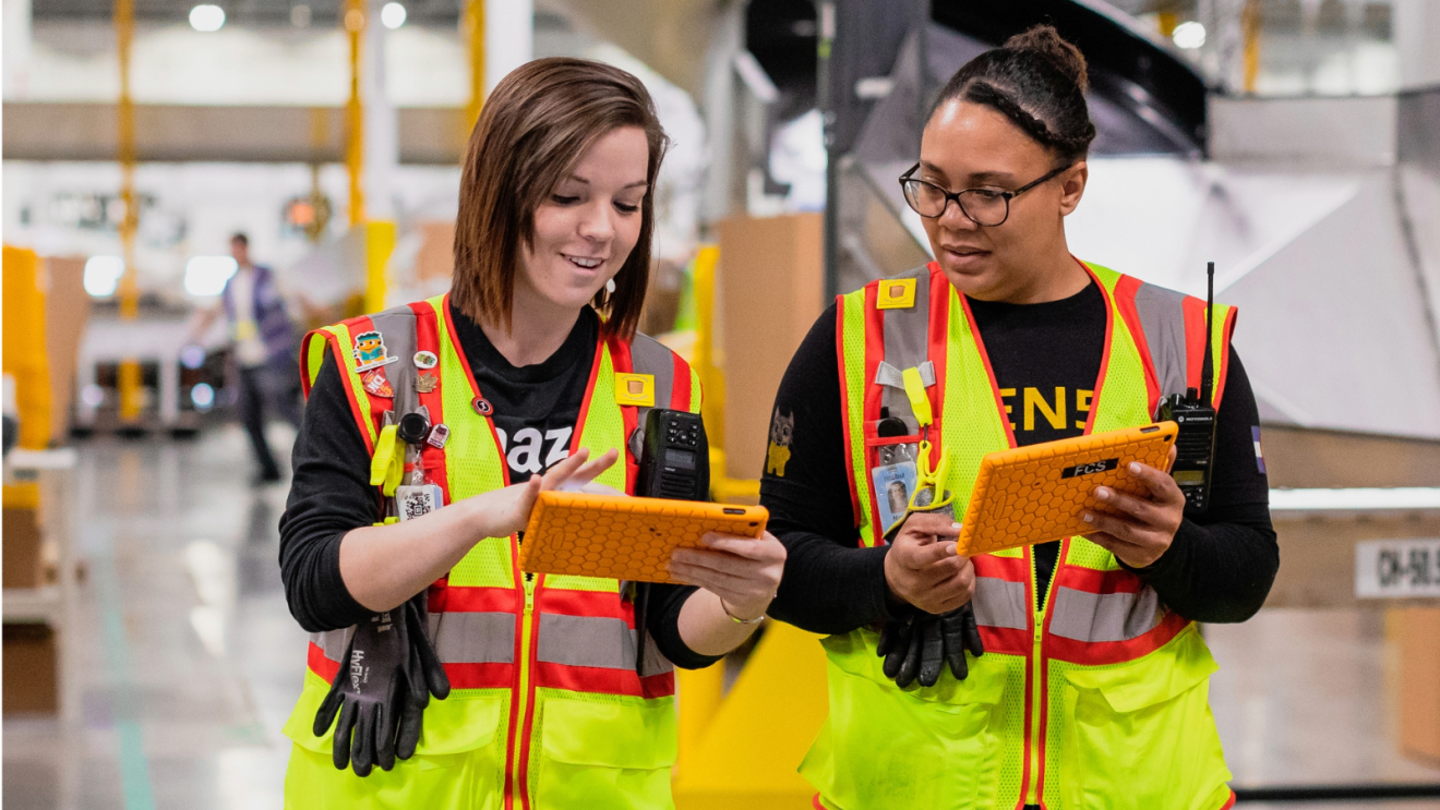 Two Amazon Employees sharing information from their tablets at work.