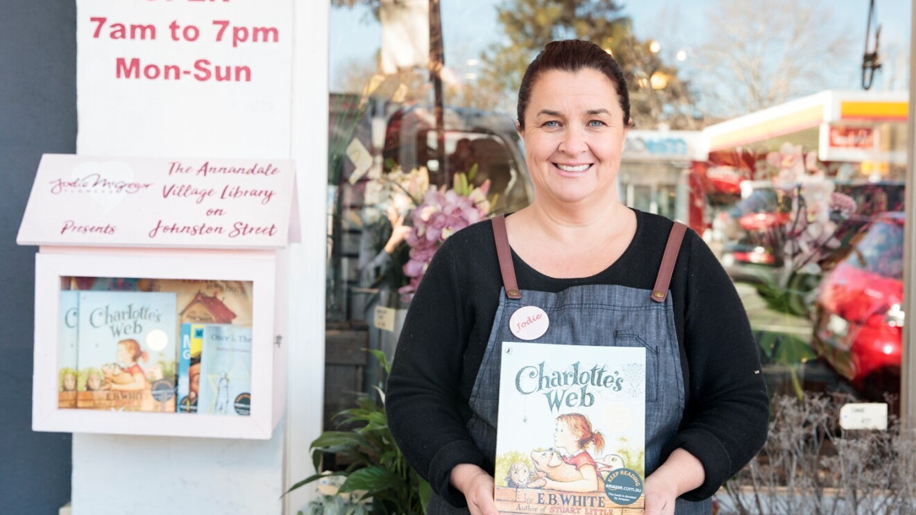 An image of a woman holding the book "Charlotte's Web," while standing in front of a storefront that has a box on its door with books available for the community to borrow. 