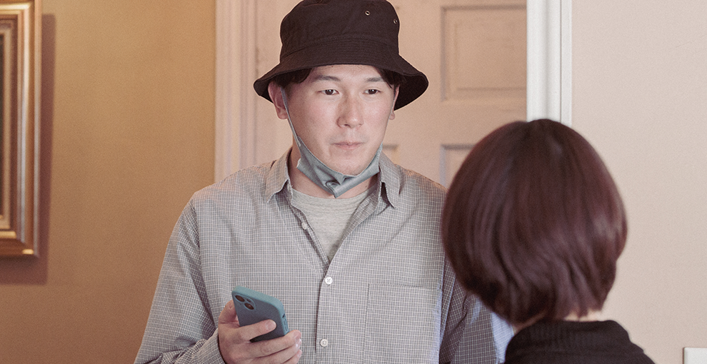 室内で向かい合う2人の人物。左側の人物は帽子をかぶり、マスクを顎にかけ、スマホを手に持っている。右側の人物は後頭部だけが見える。