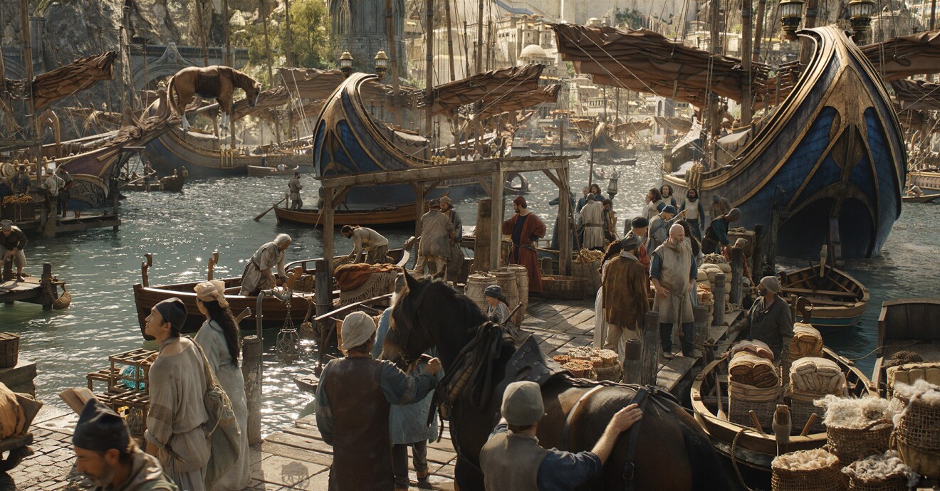 A scene from the Lord of the Rings: The Rings of Power. Showing a busy port by the sea. People are working on the land and ships are floating in the water.