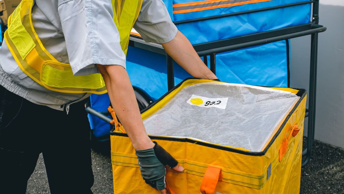 黄色い安全用ベストを着た人が緑のグローブをつけて大きな黄色い箱を運んでいる。その人の後ろには青いカバーが付いたリヤカーが置かれている。