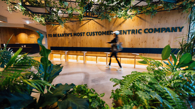 たくさんの観葉植物が置かれたオフィスの入り口。キャップをかぶった人物が歩いている。ウッディな壁には大きなアルファベット文字が設置され文になっている