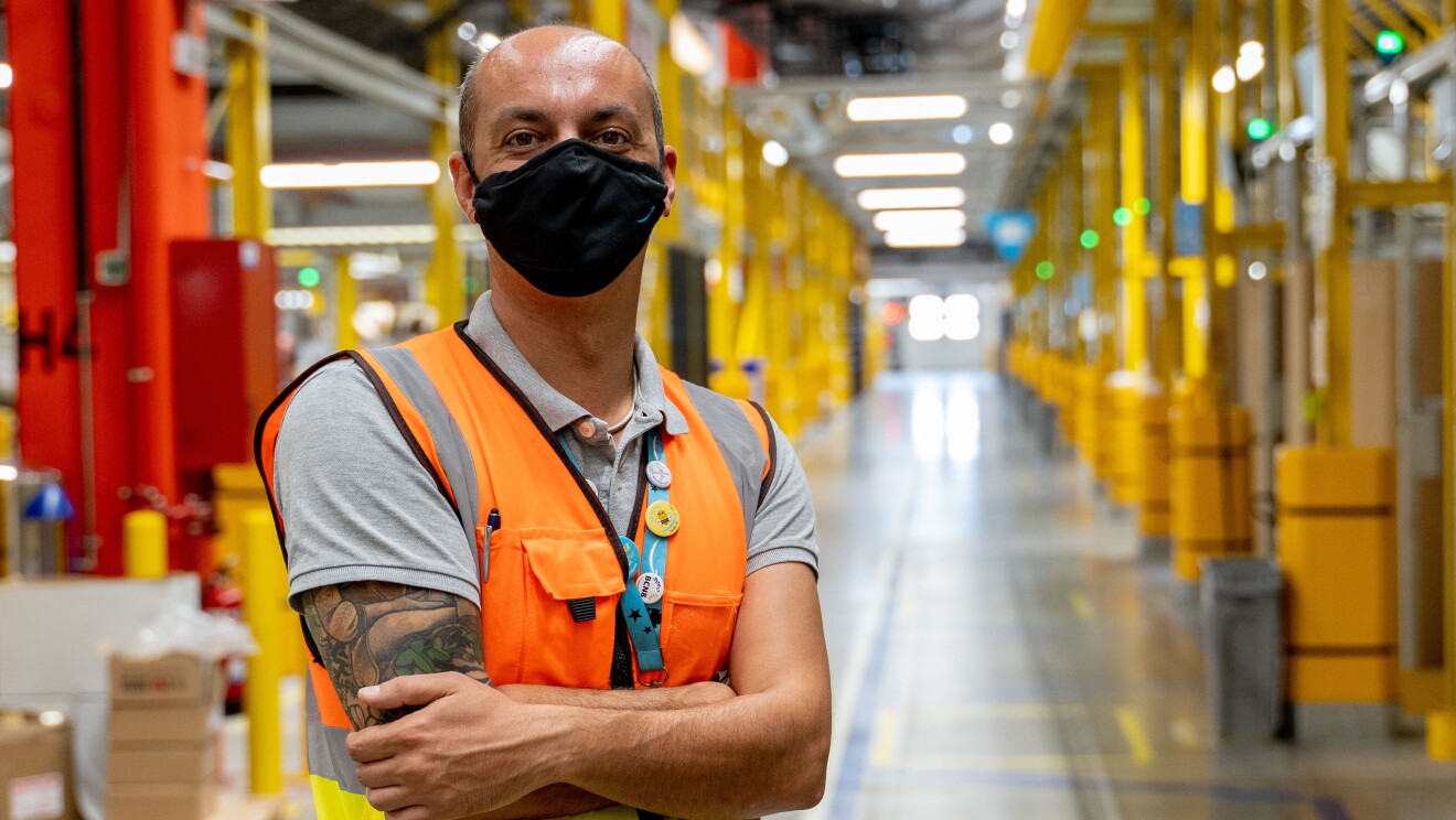 Oreste Tagliaferri, director general de los centros de distribución de España e Italia. En un pasillo del centro logístico está con los brazos cruzados Oreste. El pasillo tiene estanterías metálicas de color amarillo. Oreste tiene el brazo derecho tatuado. tiene el pelo muy corto y la parte superior no tiene pelo. Lleva una mascarilla de Amazon de color negro, un chaleco de color naranja y un polo de color gris. 