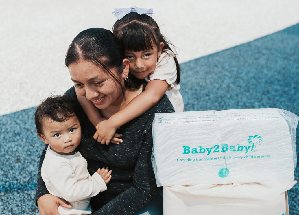 A photo of a mother holding two children, standing next to a package of diapers provided by Baby2Baby.