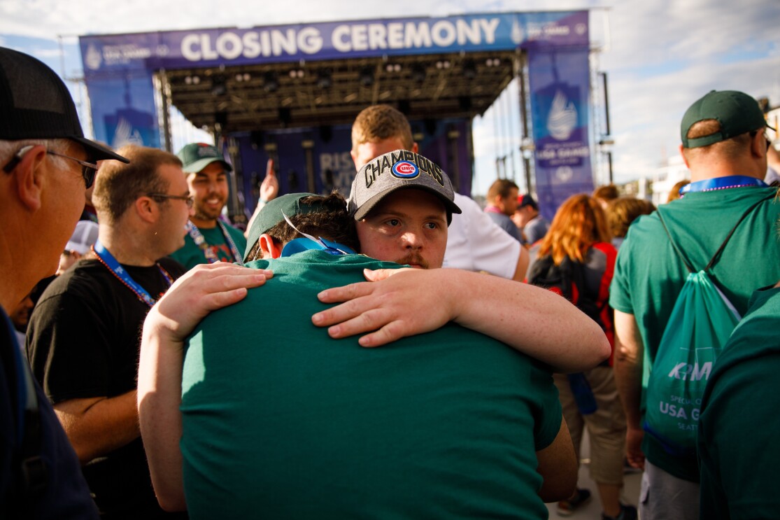 A man embraces another male, while celebrating the USA Games closing ceremony. In the background, more event attendees appear to celebrate, with a stage setup in the distance.