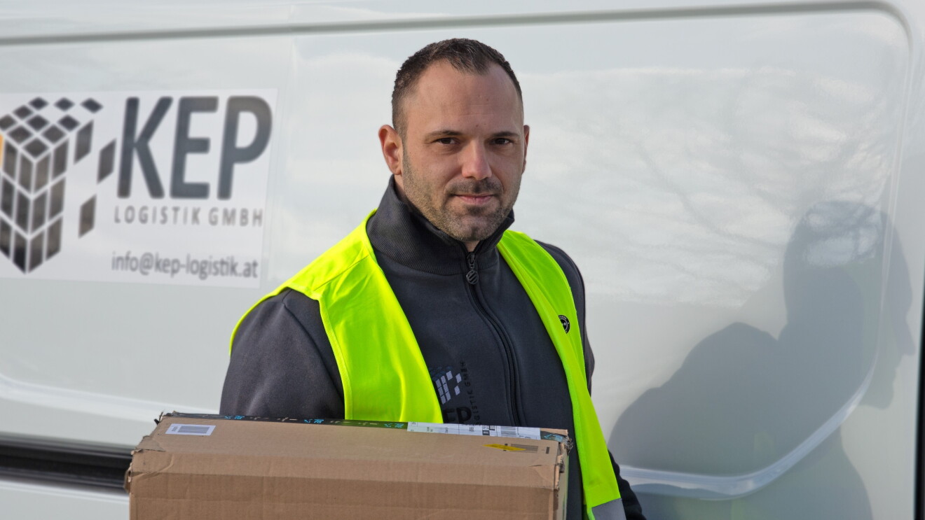 Ein Mann mit Bart in gelber Sicherheitsweste steht vor einem weißen Liefer-Van mit Aufdruck KEP Logistik GmbH. Er hält ein Paket in der Hand.