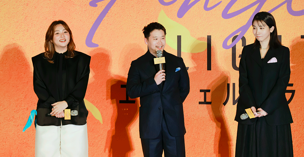 オレンジ色の壁の前、オレンジのステージの上に、黒い服を着て横一列に並んだ3人の人物。全員マイクを手に持ち、中央の人物がマイクで話している。