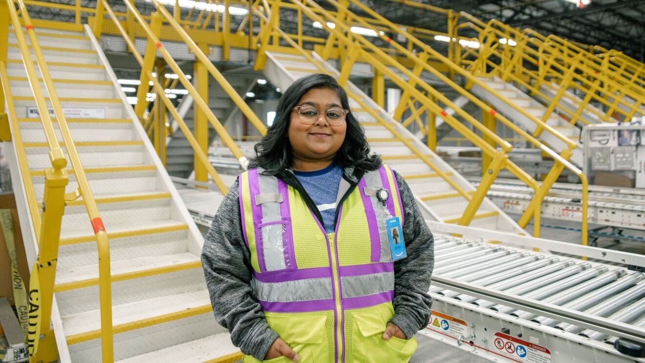 Amazon employee stands smiling with her hands in her pocket in front of a conveyor built in a fulfillment center.