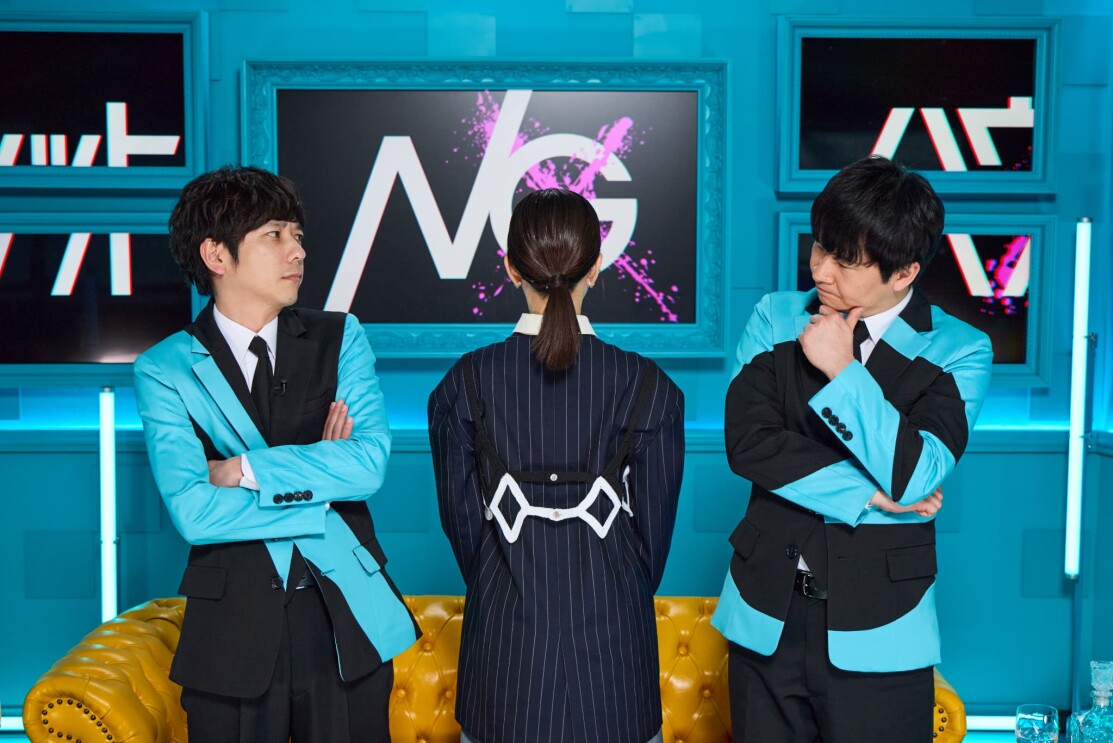 黄色のソファーが置かれている水色の部屋で、水色と黒のジャケットを着て、腕を組んで考え込む表情の男性2人（二宮和也と若林正恭）が彼らの間に立っている紺色のジャケットを着た後ろ姿の女性を見ている。背景の中央の額縁の中のスクリーンには黒地に白でNGの文字