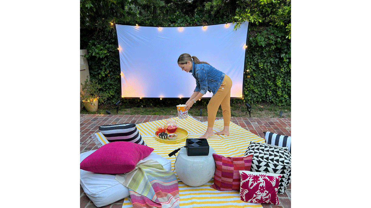 An image of Sabrina Soto in a backyard putting a bucket of popcorn on the floor. There are throw pillows in the bottom and a projecting screen in the background. There's a Fire TV stick plugged to a projector in the center.
