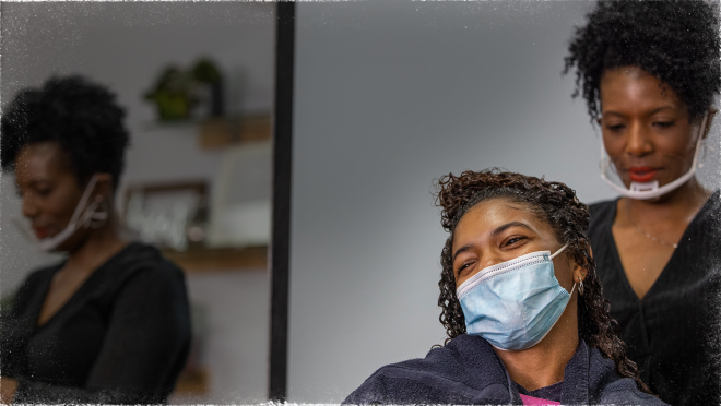 一名女子戴着口罩坐在沙龙的造型师椅子上。在她身后,另一个戴着面具的女人正在给第一个女人做发型。