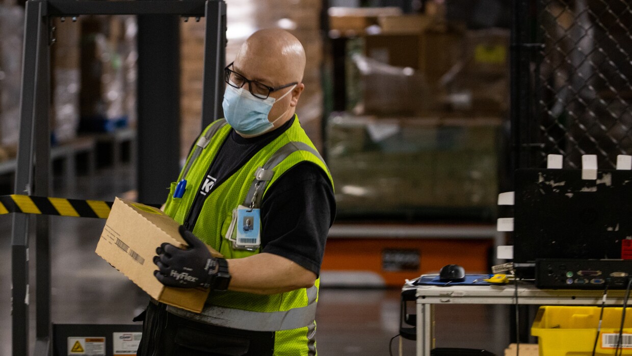 A man wearing gloves, a mask, and a safety vest carries a small box.