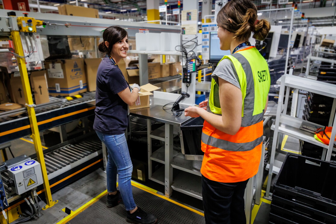 Ramona Moraru, supervisora de operaciones en la estación logística de Getafe. En el exterior del centro logístico con dos camiones de Amazon Prime de color azul con la sonrisa de Amazon en blanco. Ramona está hablando por un walkie talkie. Tiene el pelo claro, recogido en una cola, Con la mano derecha agarra el walkie talkie, tiene las uñas pintadas de negro. Lleva un tatuaje en la muñeca izquierda y un pulsera. Los ojos oscuros y maquillados y una mascarilla negra de Amazon. Lleva un chaleco de color naranja y un jersey negro con estrellas de color gris.  