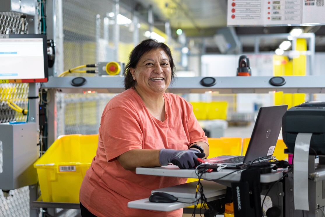 A woman stands at a computer terminal. She's wearing a tee shirt and work gloves, and leaning on the desktop. Behind her are yellow totes holding merchandise.