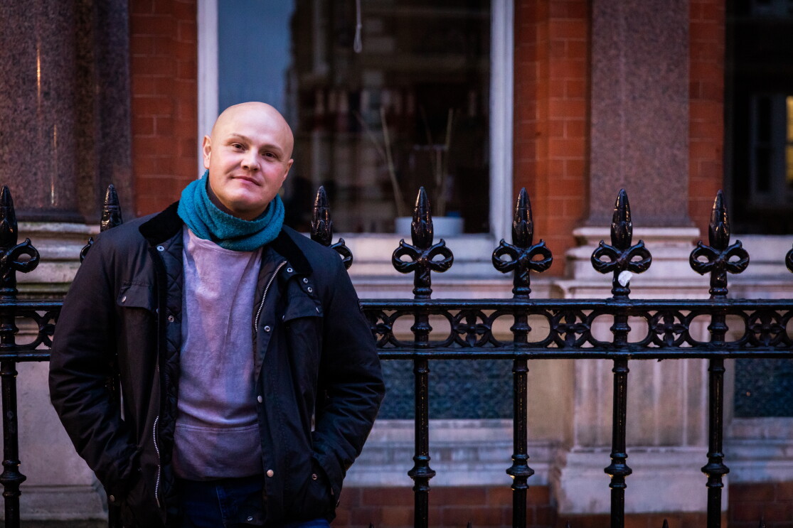 Man wearing black jacket with hands in pockets stood against some black railngs