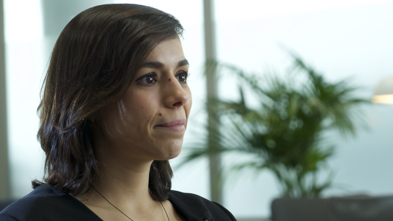 Natalia Gutiérrez, Líder Global de Impacto Sostenible y Seguridad de Cabify. De fondo unas butacas de color gris con plantas verdes y una lámpara de pie de color blanco. Unos ventanales grises grandes. La protagonista en primer plano con una camisa cruzada negra de cuello pico, manga larga y botones de color blanco. Con el pelo castaño, liso hasta el hombro y sonriendo mirando a la derecha de la foto. Lleva un collar de alambre con una bola.