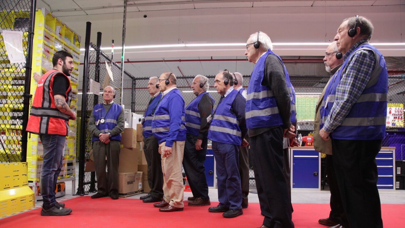 Nueve ingenieros jubilados durante la visita a un centro logístico. Todos ellos visten con un chaleco y escuchan atentamente las explicaciones del ingeniero que lleva un chaleco rojo.