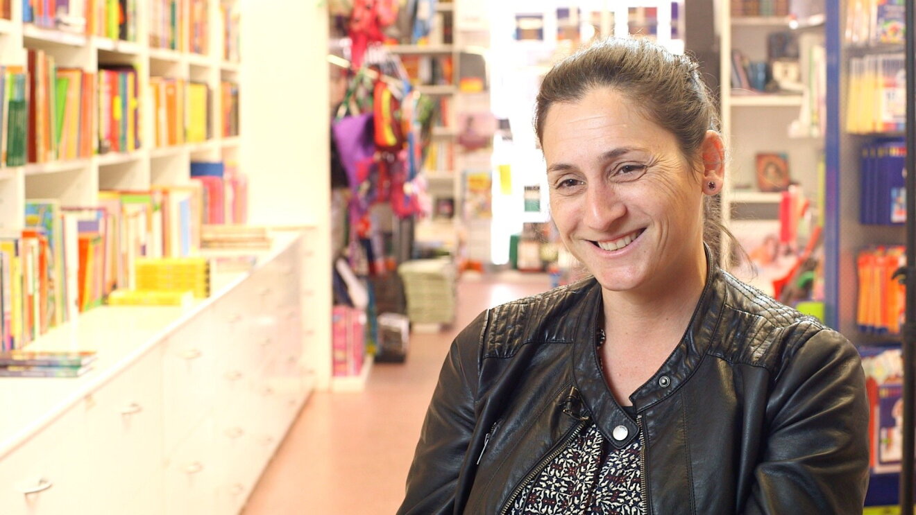 Patrícia Serrat de Llibres Text sonríe delante de dos estanterías llenas de libros. Viste con una chaqueta de cuero y un jersey negro y blanco. 
