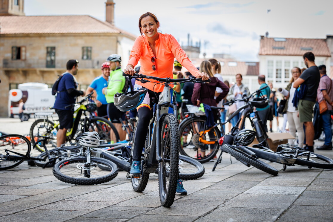 Ana Grima, trabajadora del centro logístico de Alcalá de Henares. En la plaza del Obradoiro de Santiago de Compostela está Ana encima de una bicicleta de montaña. Apoya las manos en el manillar donde tiene el casco. Va con una camiseta de color naranja, pantalones largos y unas zapatillas de color azul. Lleva el pelo recogido con una coleta. Detrás de ella hay unas cuatro bicis en el suelo y gente de pie hablando.