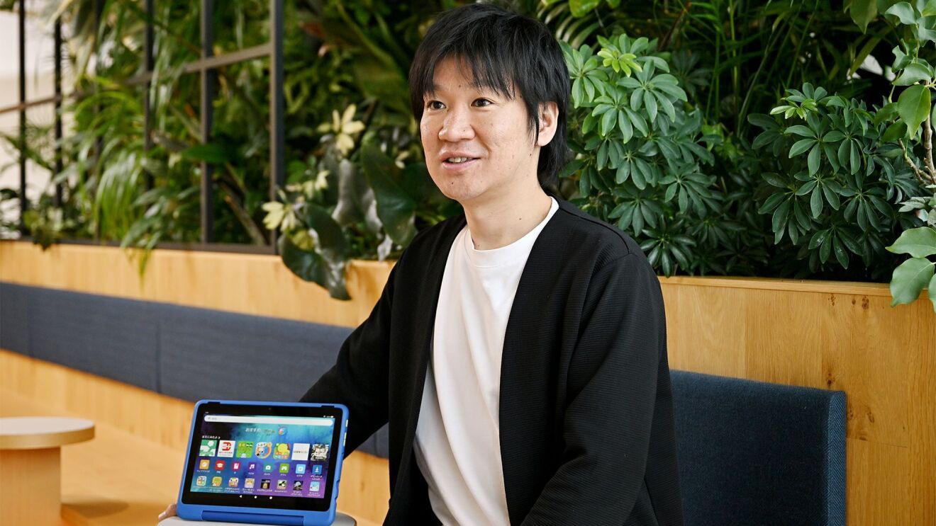 白いシャツと黒いジャケットを着ている男性が話をしている。その横に青いカバーがついやタブレットが置かれている