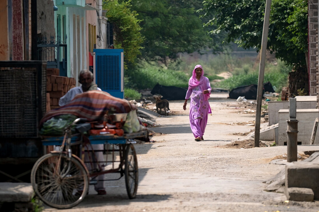 A woman walks along a dirt road. Buildings are on the left side of the image. In the foreground, a man stands behind a three-wheel bicycle loaded with cargo.