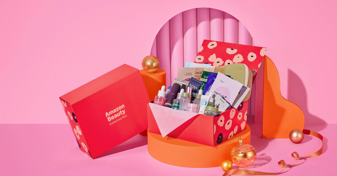 ピンク色の背景、オレンジ色台座に置かれたピンク地に花柄のボックス。蓋が開いて中にたくさんの基礎化粧商品がつまっている。左側の外箱のピンク面に「Amazon Beauty」の文字。